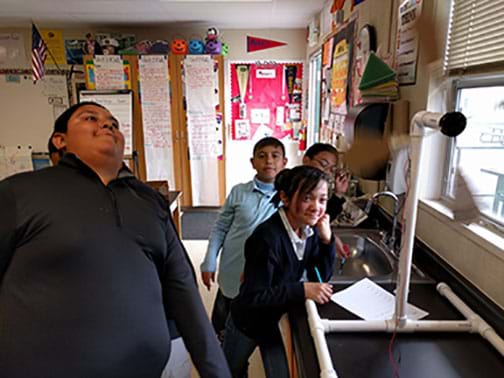 A photograph shows four smiling youngsters standing at the side of a counter in a classroom where a model wind turbine is set up and spinning, its blades a blur.