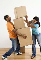 A photograph shows a man precariously carrying two large boxes while a woman places a third box on top of them.