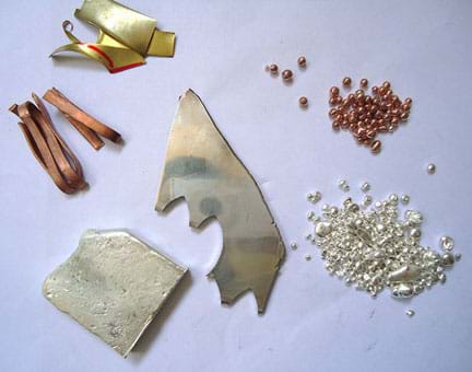 A photograph of assorted metals used for creating jewelry: scrap pieces of copper and brass, silver ingot, silver sheet, copper and germanium master alloy, fine silver granules.