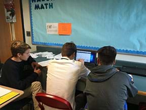 Three male students sit at a counter looking at a laptop screen showing the Desmos online graphing app.