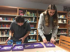 Two students are standing behind a table with six purple rectangular shallow bins containing stickers.  The student on the left is looking at the stickers they have placed in a giant plastic egg; the student on the right is using her hands to search for particular stickers.