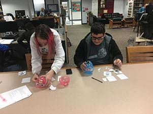 Two students are affixing face feature stickers onto large plastic Easter eggs.  A female student is standing and placing stickers onto a pink egg; a male student is sitting and placing stickers onto a blue plastic egg.