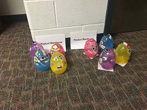 Two signs entitled “Product Warehouse” are displayed.  In front of each sign are four plastic Easter eggs with face feature stickers affixed.  There is a purple, blue, yellow, and pink egg in each grouping.