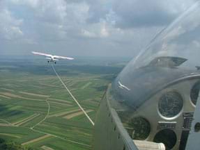 A photograph shows the view from inside an airborne glider: a glass-enclosed instrument panel, a rope connected to a small plane flying ahead of it, and green fields and blue sky beyond.