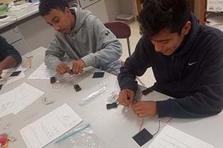 A photograph shows two teen boys sitting at a table, working with wires as they build their solar USB chargers.