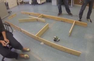 A photograph shows the legs of three teens watching and controlling a wheeled robot on a floor area of tiled linoleum to move through a maze made from long wooden boards positioned to stand on their thin edges to create low walls.