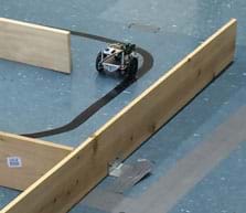 A photograph shows a wheeled robot on the floor navigating a maze made of wooden plank walls.