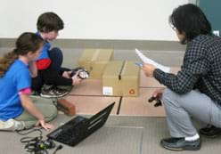 A photograph shows a teacher and two students around a maze on the floor made of boxes and tape lines. They are using a laptop and LEGO robot.