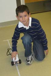 A photograph shows a young boy holding his LEGO robot on a tape measure line on the floor.