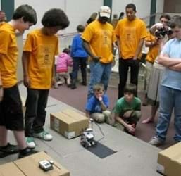 Students and teachers gather around a LEGO robot on a classroom floor along with various cardboard boxes.