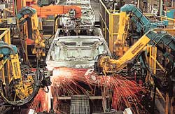 Photo shows car chassis moving through an automated assembly line, surrounded by robots performing welding and other tasks.