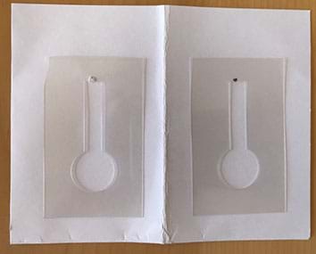 Photograph of a white card that has been folded down the middle and opened up to the inside. Two stencils cut from transparent acetate have been laid on either side.  Each stencil consists of a rectangle with a thermometer-like shape cut out: a long stem with a round bulb on the bottom.