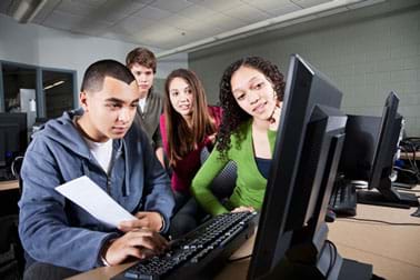 A photograph shows four teens around a computer, looking at the monitor and working on a project.