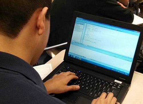 A photograph from behind a boy working at a laptop analyzing lines of computer code.