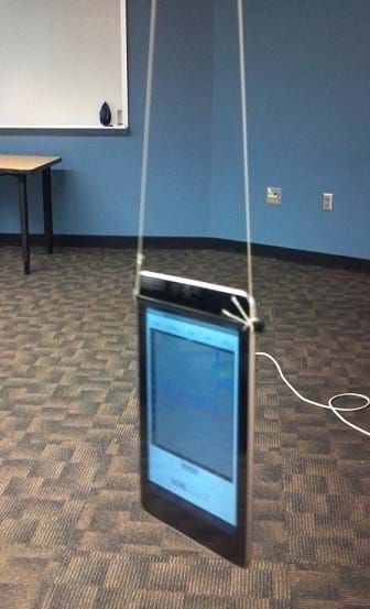 A photograph shows a seven-inch Android tablet computer hanging from a classroom ceiling via two strings attached at the top left and top right of the flatscreen device.