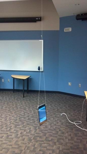 A photograph shows an Android tablet computer hanging from a classroom ceiling via two strings attached at the top left and top right of the flatscreen device. The tablet serves as a pendulum weight.