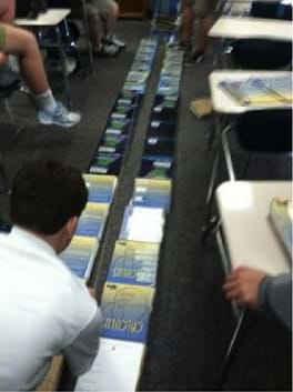 Photo shows students on the floor with two long lines of textbooks about 10 cms apart. A student in the foreground holds one end of a rubber band string that runs the length of the textbook "track."