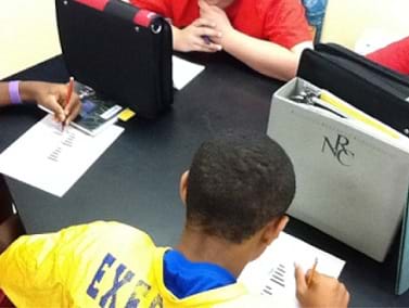Photo shows students playing the game of battleship. Students have a sheet of paper in front of them and sit facing another student. Binders are placed on the table, standing vertically, between the students, so that students cannot see any other student's sheet of paper.