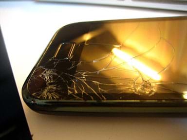 A photograph shows an iPhone with a cracked screen on a table with a ceiling lamp reflected in the glass. 