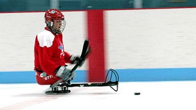 Sled hockey player on sled using hockey sticks to maneuver on the ice towards the puck.
