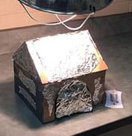 A photograph shows an illuminated clamp lamp positioned above what looks like a small, foil-covered cardboard peaked-roof house sitting on a laminate countertop. Nearby sits a thermometer.
