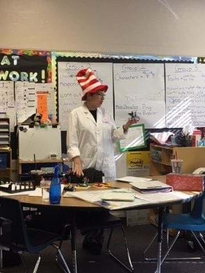 A teacher stands behind a table of supplies, wearing a lab coat and a Dr. Seuss “Cat in the Hat” hat. She is holding her Lego kingdom. There are other Lego kingdoms on the table, along with the “rain” (A cup of water).