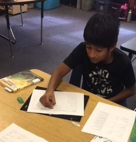 A male fifth grader sits at a table. He is writing in his design plan and has a copy of the material constraints next to him.