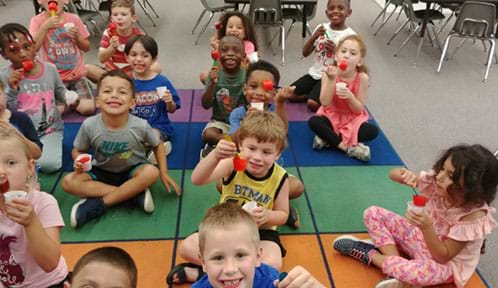 Fifteen kindergartner students sit on colorful mat eating red popsicles.