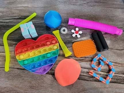 Many fidget tools sit on a table. There is a bending tube, a Pop-it shaped like a heart, a stress ball, a tactile brush, a stretch ball, and a bendy wood toy. 