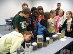 Photo shows nine kids examining 11 jars of clear and murky liquids lined up on a table.