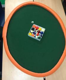 A photograph shows a view from above an elliptical-shaped pool table with a set of pool balls on a green surface material with a curved orange foam “curb” border around its perimeter. 