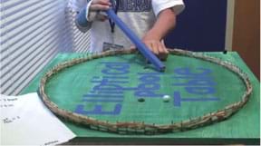 A photograph shows a student using a blue wooden stick as a cue stick to move a white marble on an elliptical pool table that is a painted wooden board enclosed by a fence (bumper edges) of rubber bands stretched on a ring of nails pounded into the perimeter.