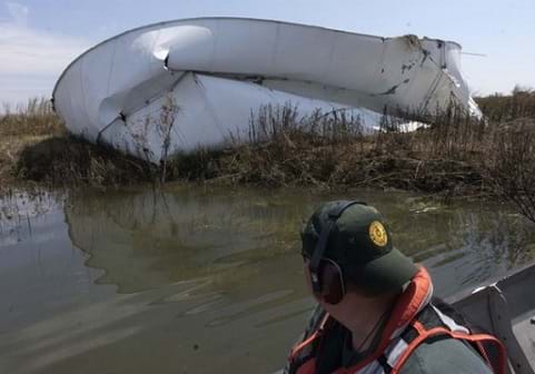 Photo shows a man in a small boat looking at a large mangled pile of white steel (a ruptured above-ground oil storage tank) in a marshy area.