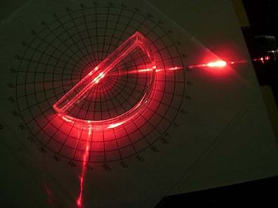 A photograph from above shows a red beam of light from a laser pointer laying on a tabletop shining at and being reflected away at a different angle from a semi-circular hollow block that sits centered over a polar graph.