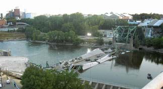 A photograph shows the scene at the I-35W Mississippi River Bridge after its August 2007 collapse. The bridge deck is in the water with a few cars on it, broken off from the rest of its steel truss components at the river’s edge.