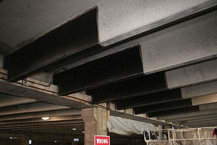 A photograph of the concrete ceiling of a parking garage shows an example of concrete structures being strengthened by CFRP reinforcement in the form of a three-sided wrap of six beams.