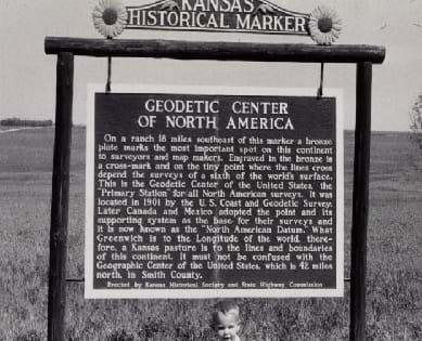 A black and white photograph displays a hanging sign in front of a an empty field. The post holding the sign reads "Kansas Historical Marker," and the main sign reads "Geodetic Center of North America."
