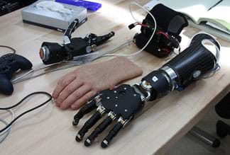 A photograph shows an assortment of parts and wires on a tabletop, including a black and silver robotic-looking forearm and hand as well as a covering for the hand that looks convincingly like a real human hand (skin, nails wrinkles, hair).