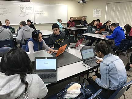 A high school classroom, with four groups with four to five students in each group, each student is working at a laptop computer, actively engaged working.