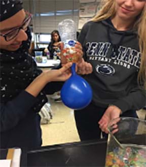 Two students working together, one holds the balloon onto a plastic water bottle, the other hold the water bottle. The students are transferring the pre-counted hydrogels from the water bottle to the balloon.