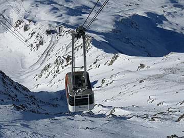 A photograph shows an aerial tram ascending above a snow-covered, rocky mountain. Far below in the distance is a docking building.  