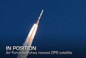 A photograph shows a blasting off rocket (equipped with a GPS satellite) as it propels through the air, trailing a stream of flame and smoke from the engines. The rocket is red on top and white on bottom with a red stripe in the middle. Overlaid text reads “IN POSITION. Air Force launches newest GPS satellite.”