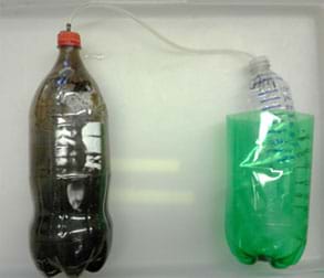 A photograph shows a capped 2-liter bottle on the left with a tube connecting to a capped, open-bottom container floating in a container of water on the right.