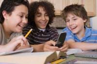A photograph shows three boys at a table with books and pencils, laughing at something they can all see on a cell phone screen.