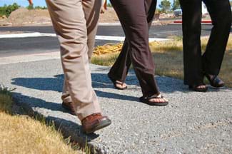 A photograph shows the legs and feet of three people walking on a gray sidewalk that looks like it is made of much aggregate, as spaces can be seen between the aggregate pieces.