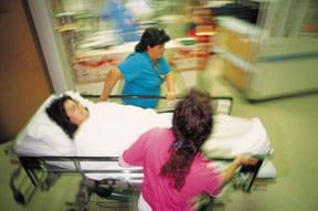 Photo shows two running hospital workers wheeling a woman on a gurney down a hallway.