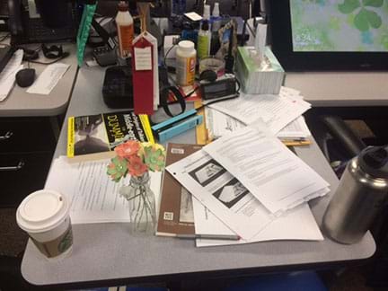A photograph shows a teacher’s classroom desk cluttered with many objects—papers, books, coffee cup, water bottle, pencils, pens, Kleenex box, vitamin bottle, lotion bottle, camera, awards, sunscreen bottle, stapler, paperclips, picture frame, flowers, etc.