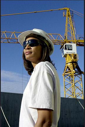 A photograph shows a woman wearing a hard hat and wrapped sun glasses at a construction site with a crane in the background.