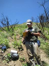 Photo shows a man standing on a dry hillside taking notes in a field book. Other equipment and materials are scattered nearby.