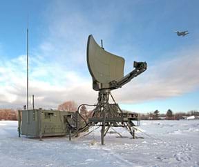 A photo shows an airplane flying over the Lockheed Martin TPS-79 mobile surveillance and air traffic control radar system, a 30-foot high piece of equipment mounted on tripod legs with an arc-shaped satellite dish panel.